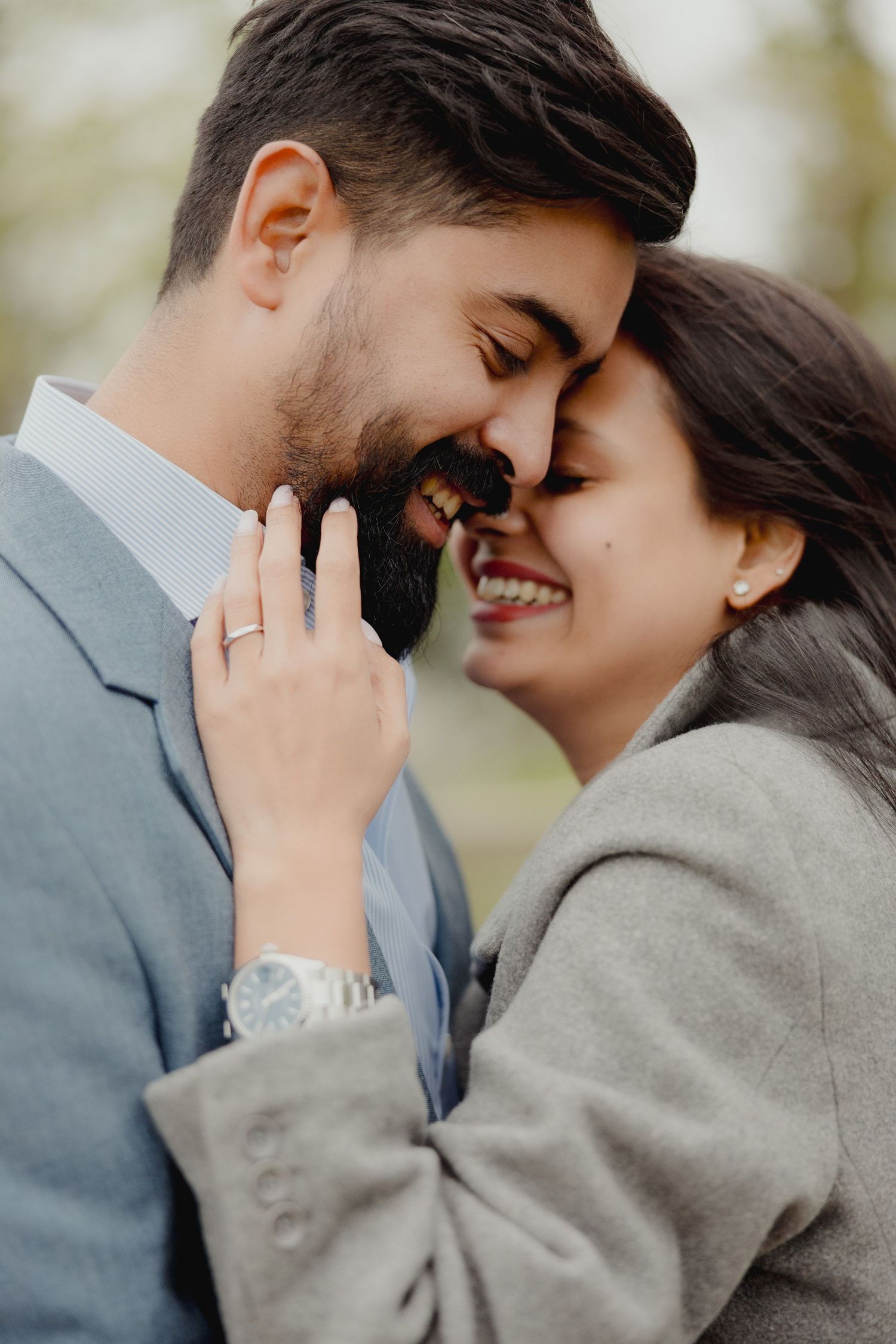 A smiling couple in gray coats share a romantic forehead touch, the woman displaying an engagement ring on her hand.