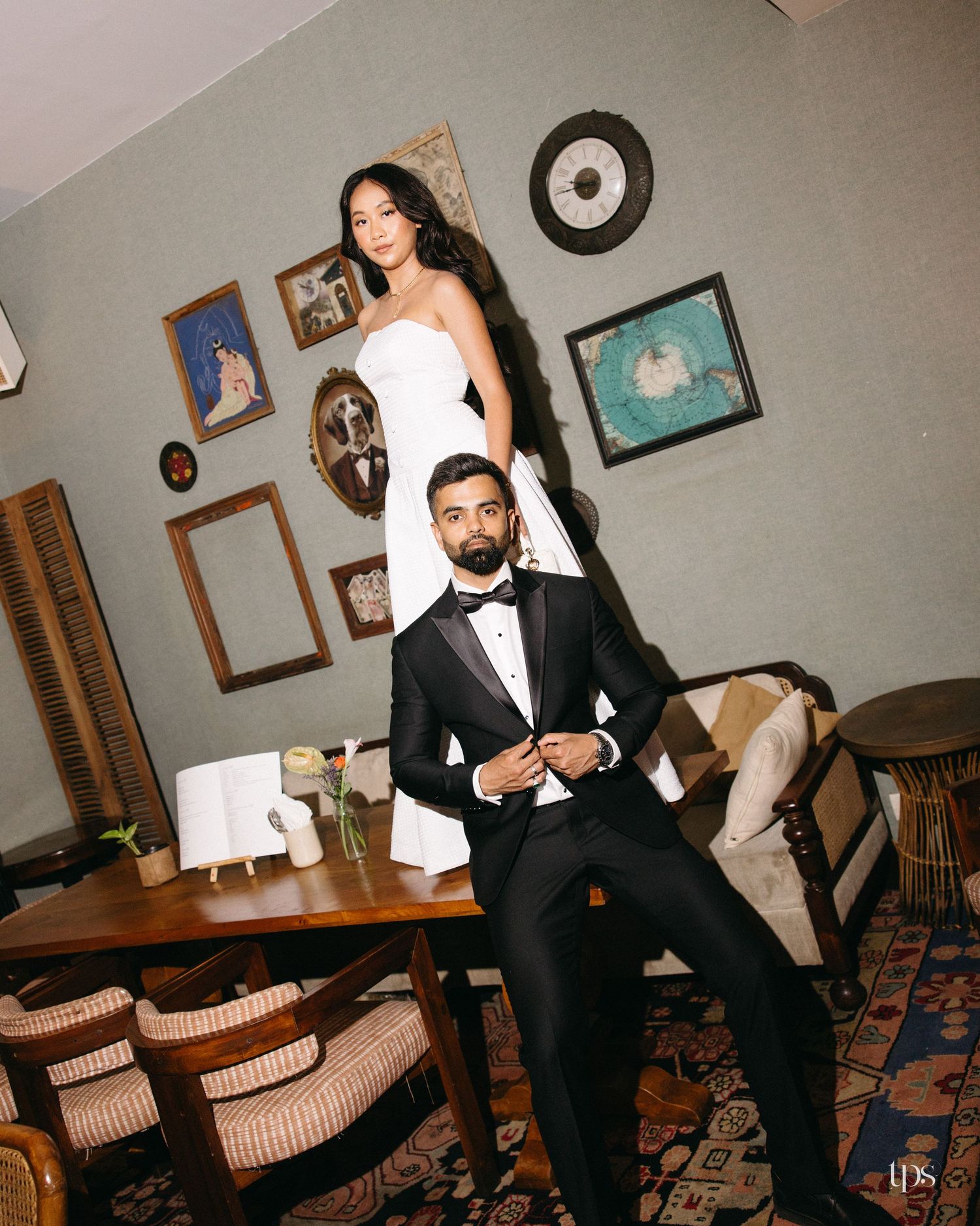 Elegant couple in wedding attire pose in a vintage-styled room, man in tuxedo seated while woman in white gown stands behind.