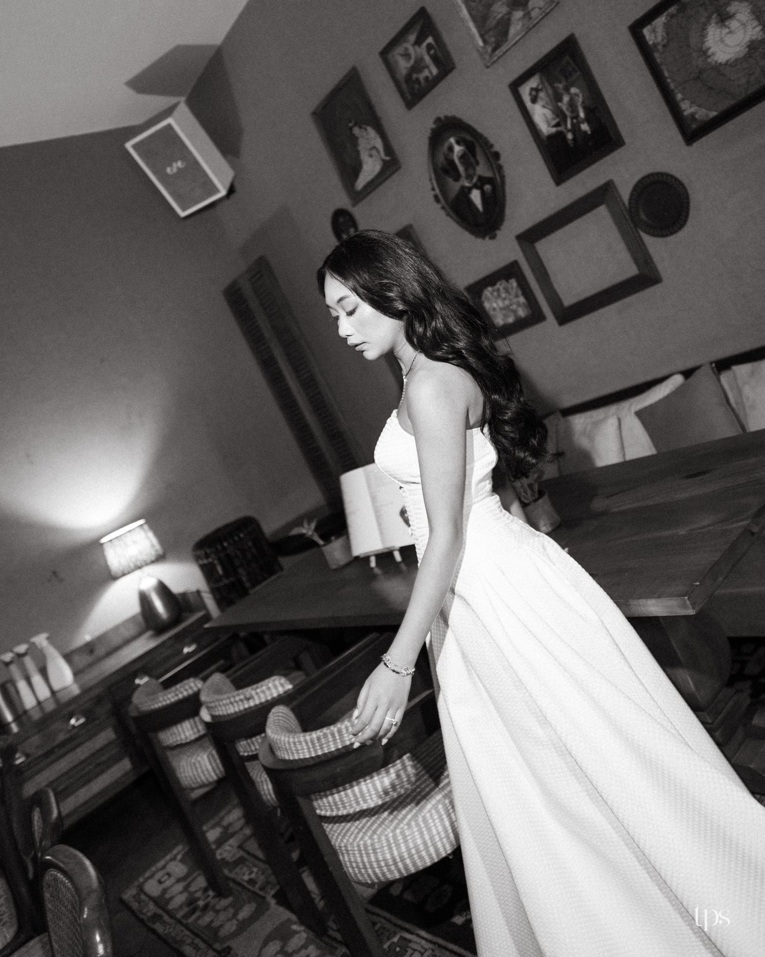 Black and white photo of elegant woman in white dress standing in a vintage room with framed wall art.