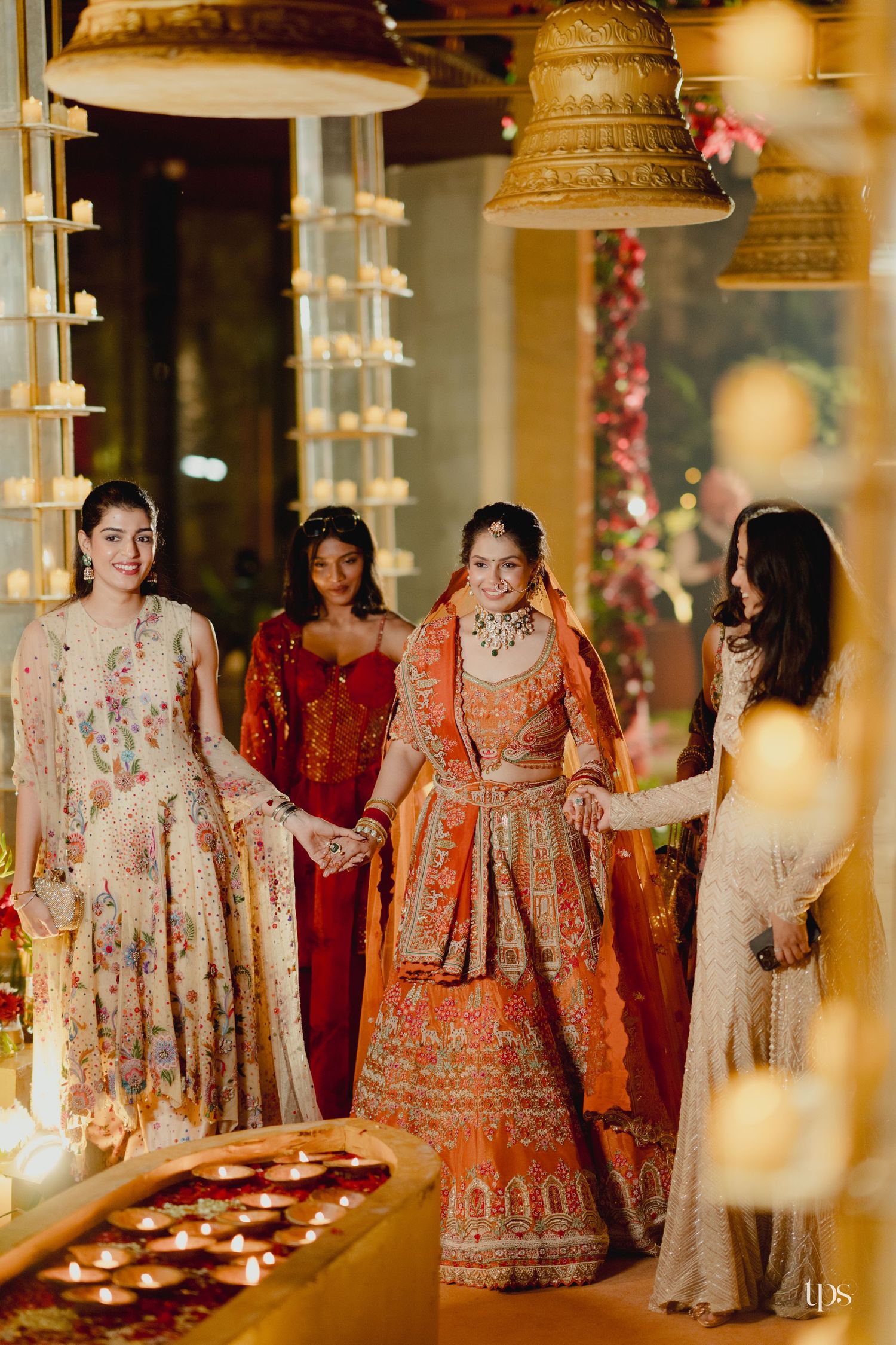 Indian bride in orange lehenga walks with bridesmaids amid glowing diyas and golden bells captured by wedding photographer in Mumbai near me