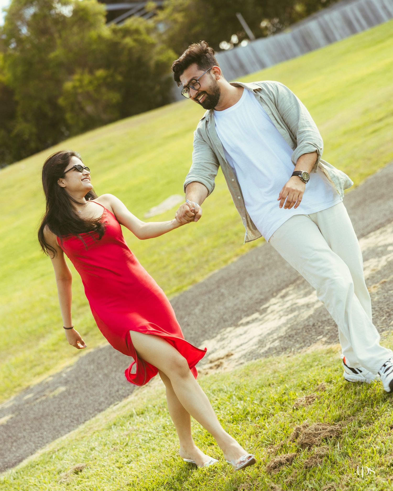 Couple dancing outdoors on grass, woman in red dress holding hands with man in white outfit and gray jacket.
