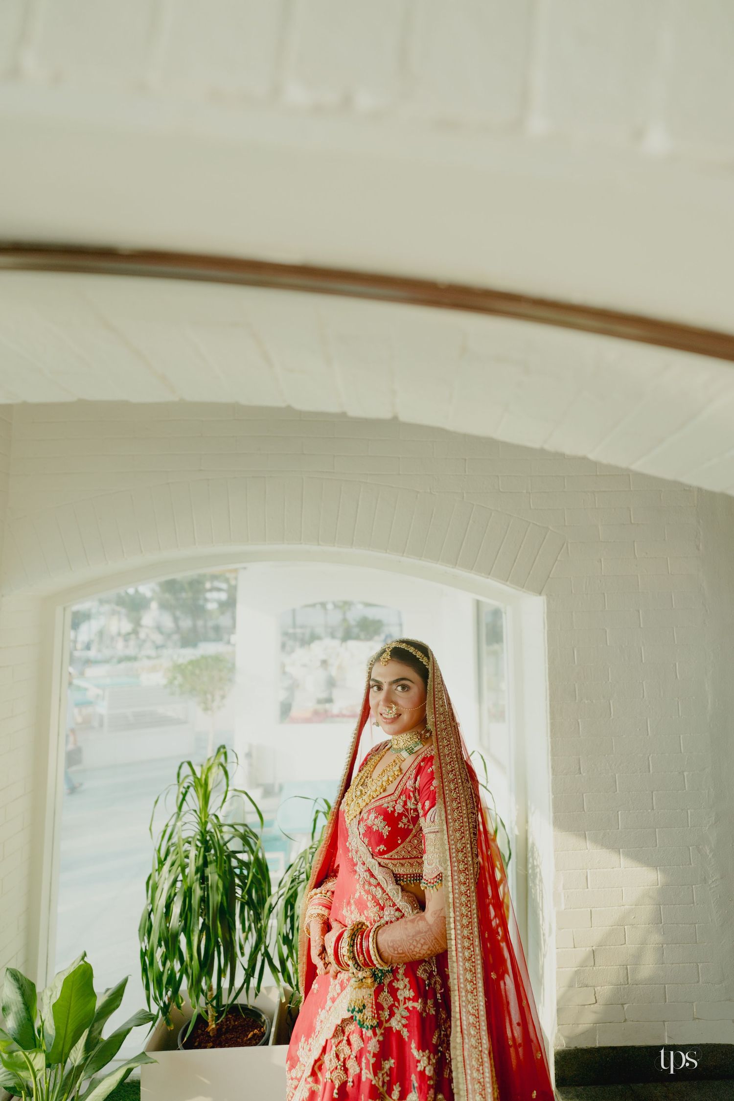 Bride in red floral lehenga with gold dupatta poses in an arched doorway by a wedding photographer near me, London & Birmingham.