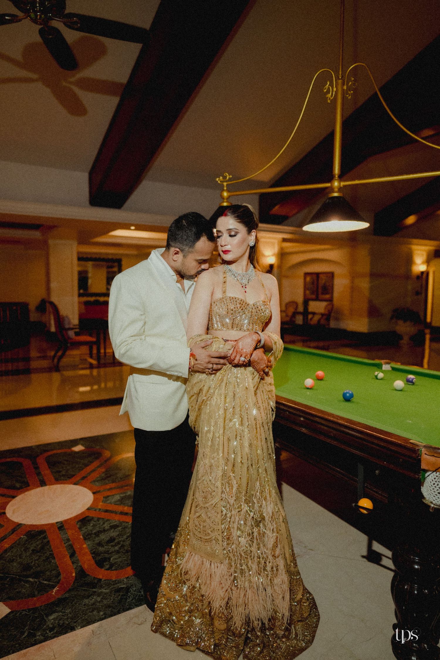 Couple in elegant wedding attire pose near a billiard table in a luxurious venue, bride in golden lehenga.