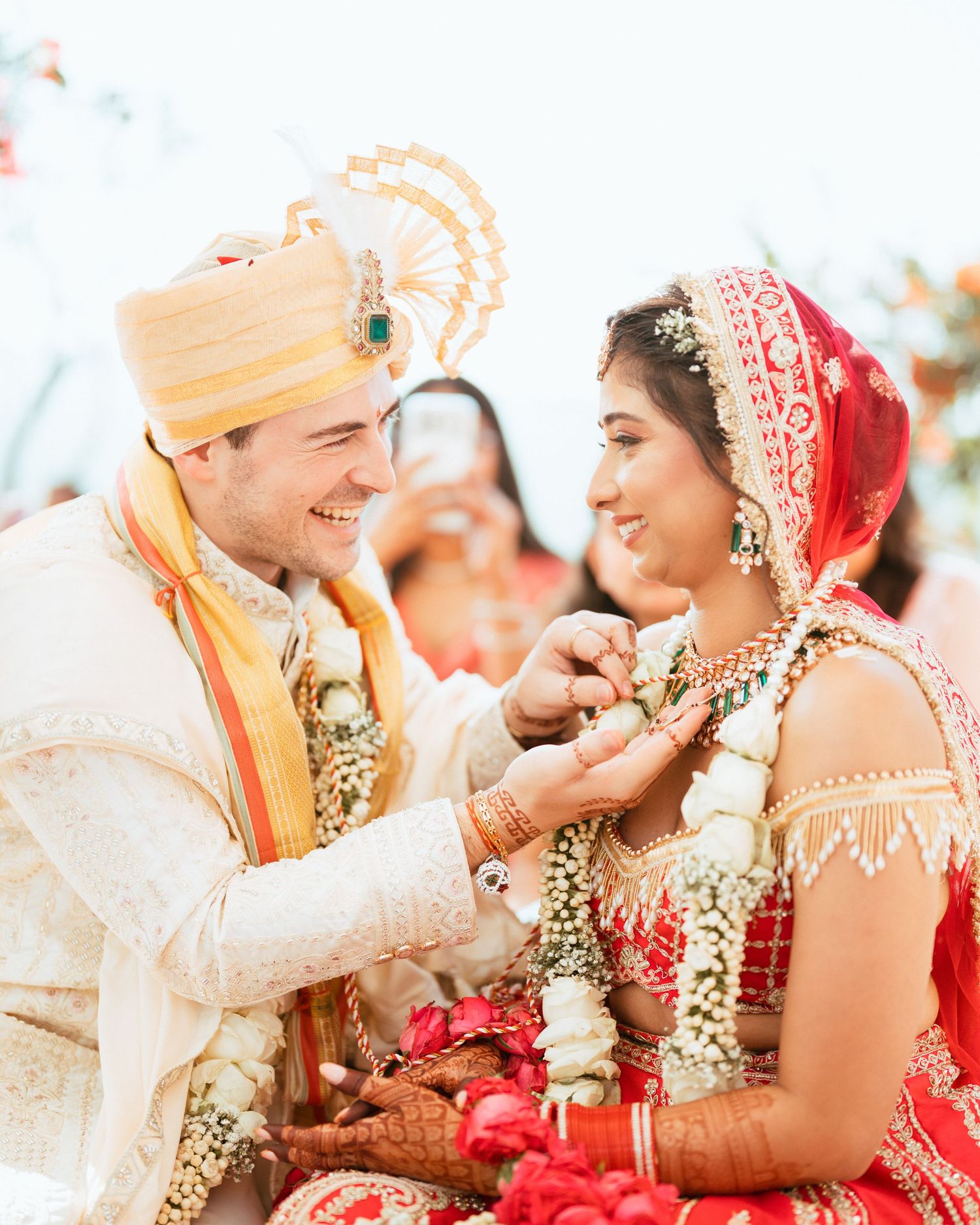 Couple smiling during traditional Indian wedding ceremony, groom in cream sherwani and turban, bride in red bridal lehenga.