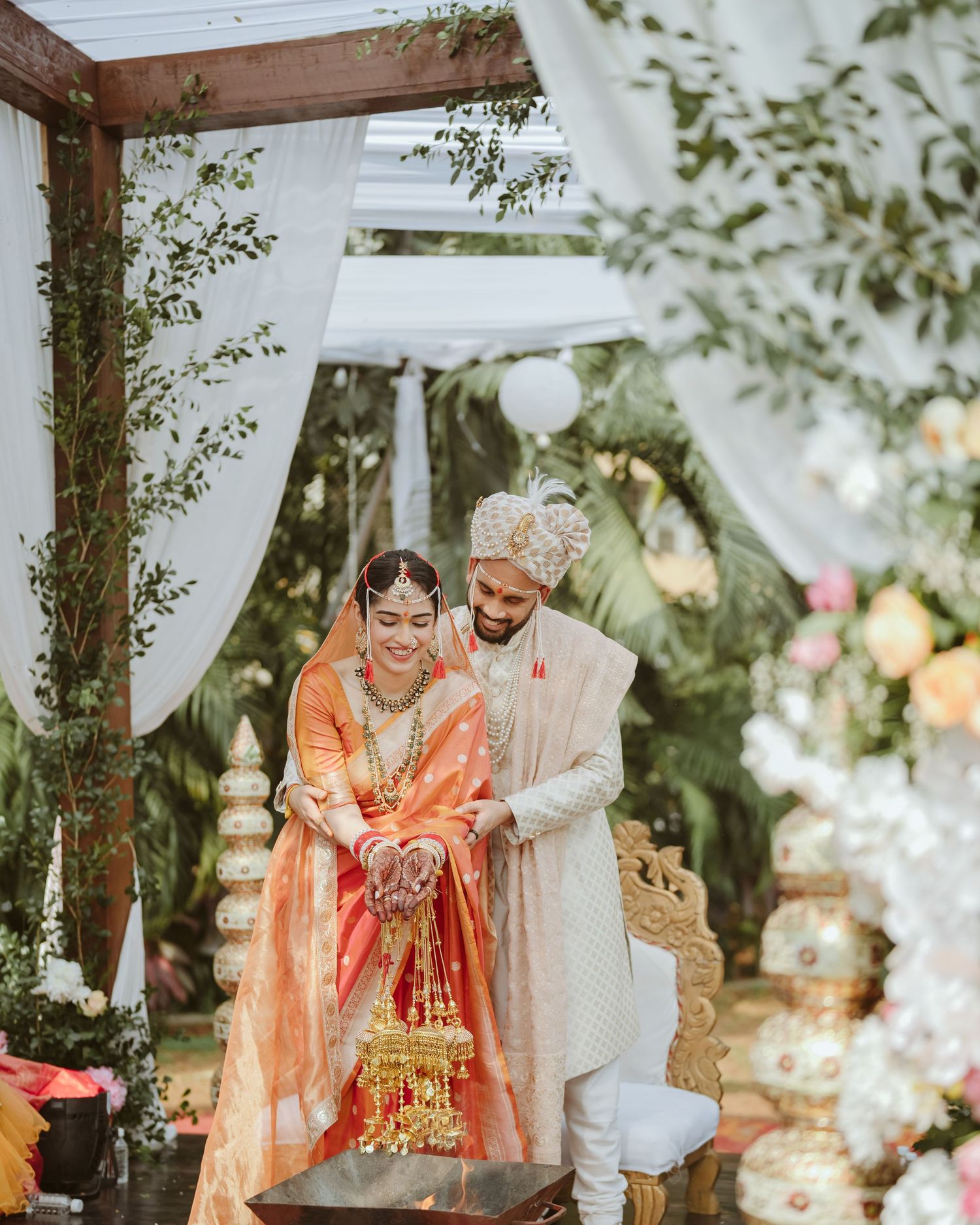 Stunning Indian wedding couple in orange & cream attire under a wooden pergola, shot by a trusted wedding photographer near me in Mumbai.