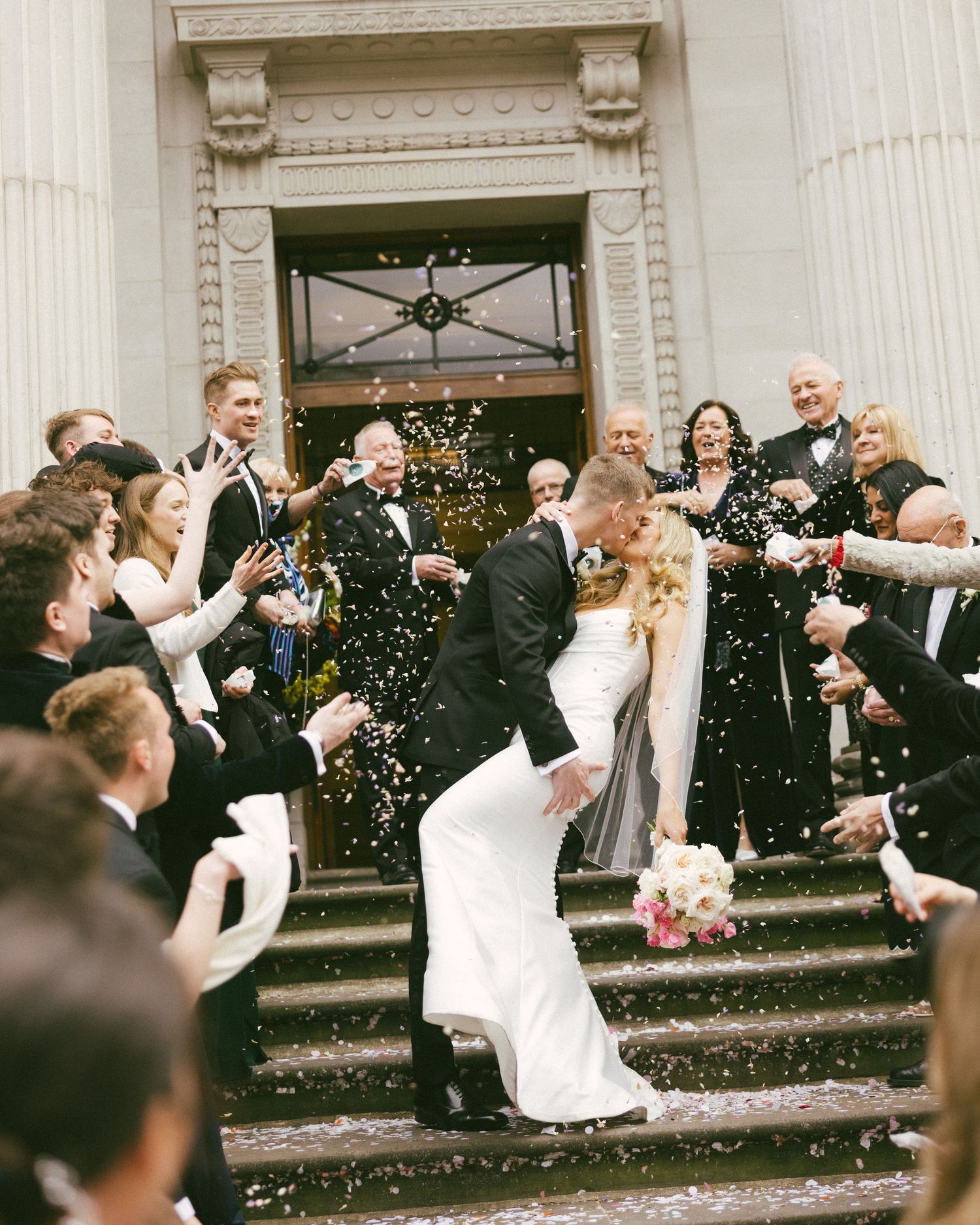 Bride and groom share a romantic dip kiss on church steps while guests shower them with confetti at their wedding.