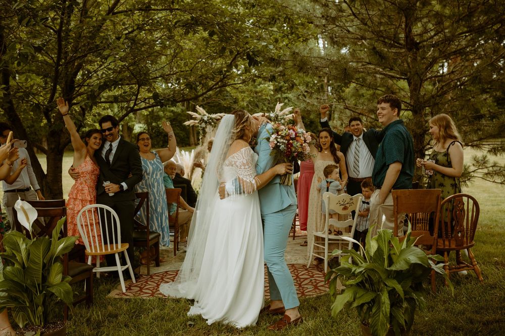 Joyful bride in white gown celebrates with partner in teal suit after outdoor garden wedding ceremony.
