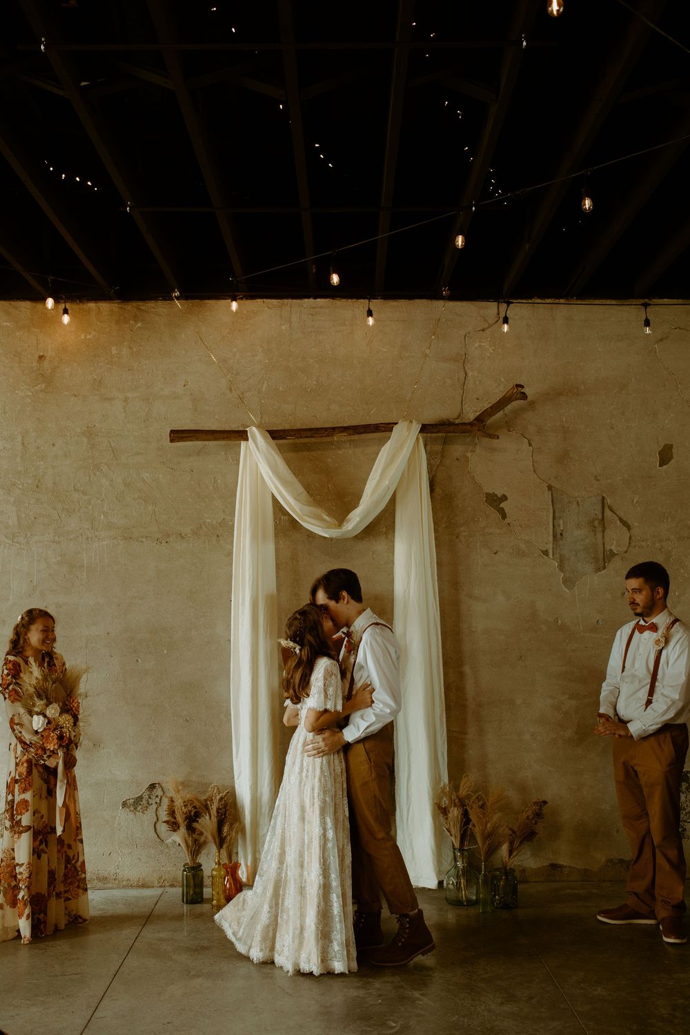 Bride and groom sharing first kiss under rustic wooden arch with white draping in industrial venue.