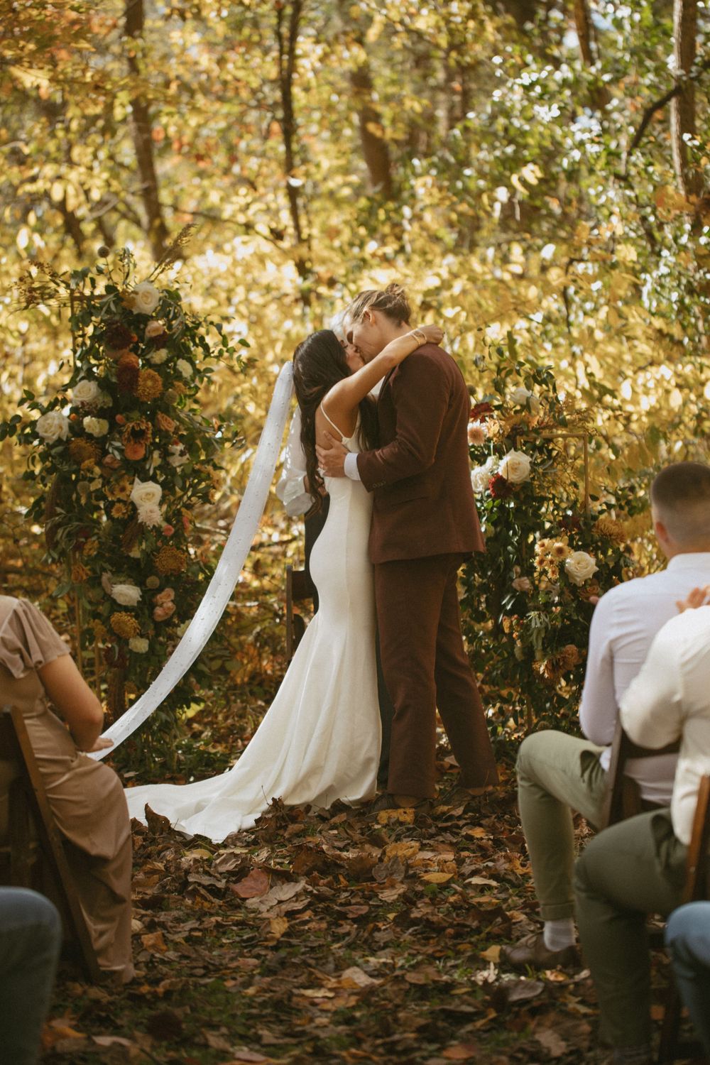 Bride and groom share first kiss at outdoor fall wedding ceremony surrounded by colorful autumn foliage.