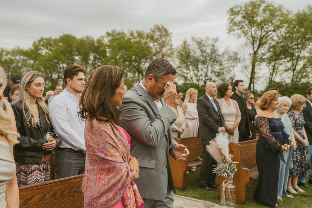 Emotional wedding guests standing in outdoor ceremony, man wiping tears while crowd watches in lush green setting.