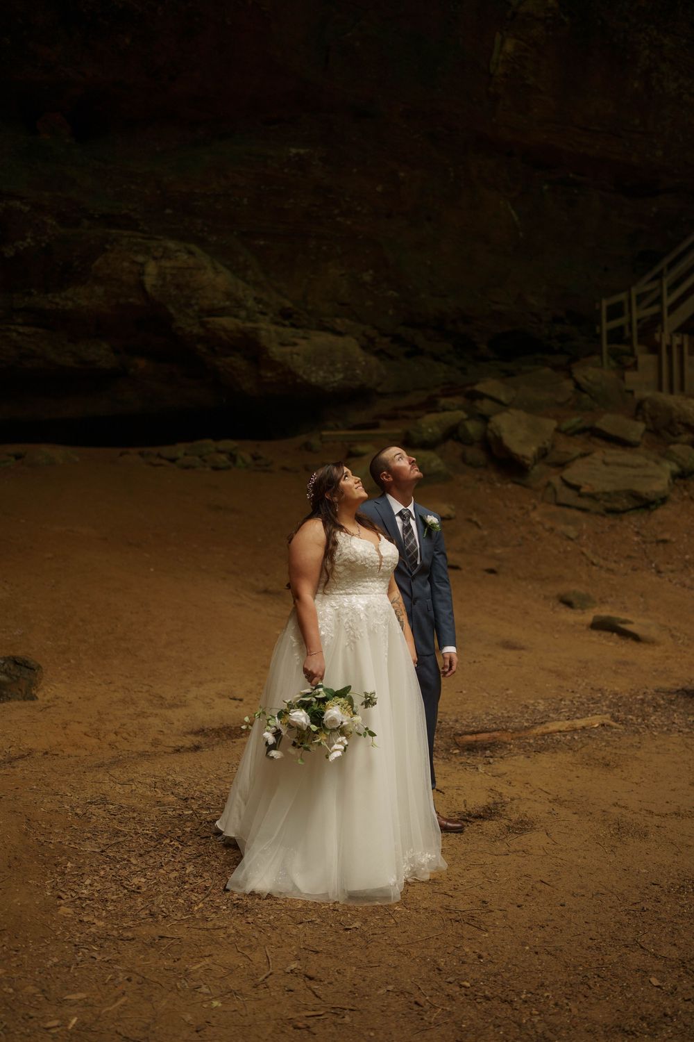 Bride and groom gazing upward inside dramatic cave, bride holding white bouquet in flowing gown.