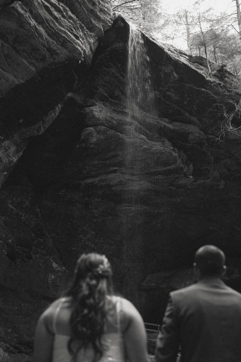 Black and white photo of bride and groom viewing waterfall cascading down rocky cliff face.