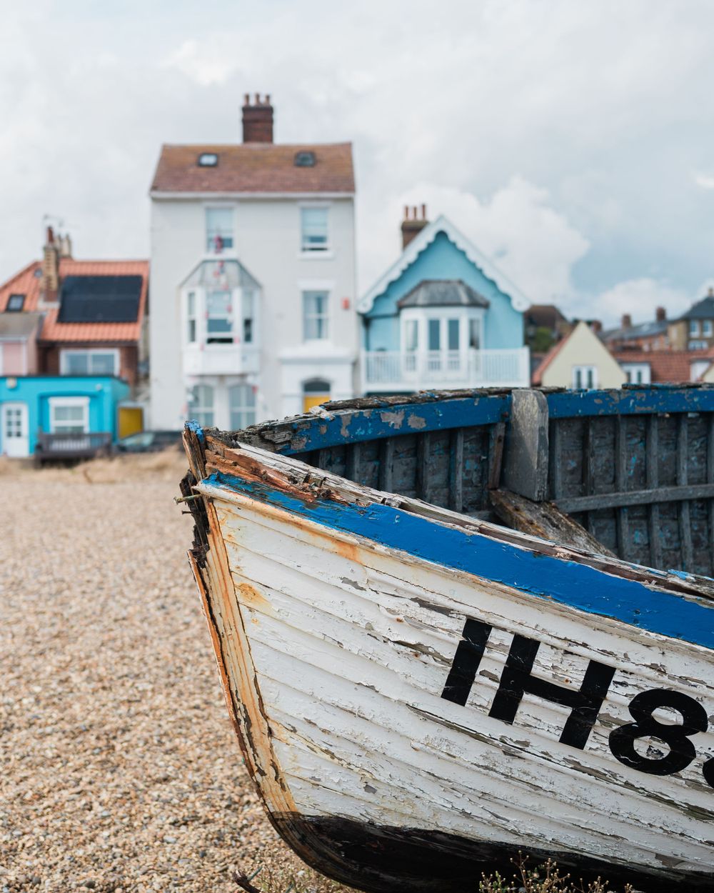 Weathered blue and white wooden fishing boat hull rests on a beach with coastal houses in the background.