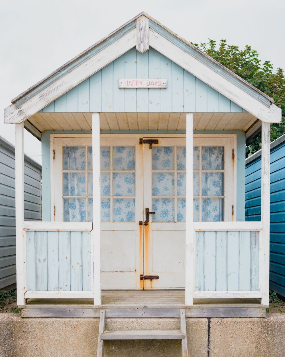 A row of vintage white wooden beach huts with blue trim and glass windows stand along a seaside promenade.