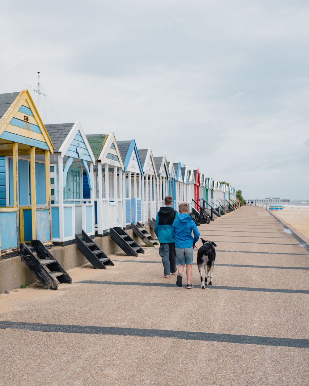 Colourful beach huts line a sandy boardwalk along the coast on a cloudy day with visitors walking past.
