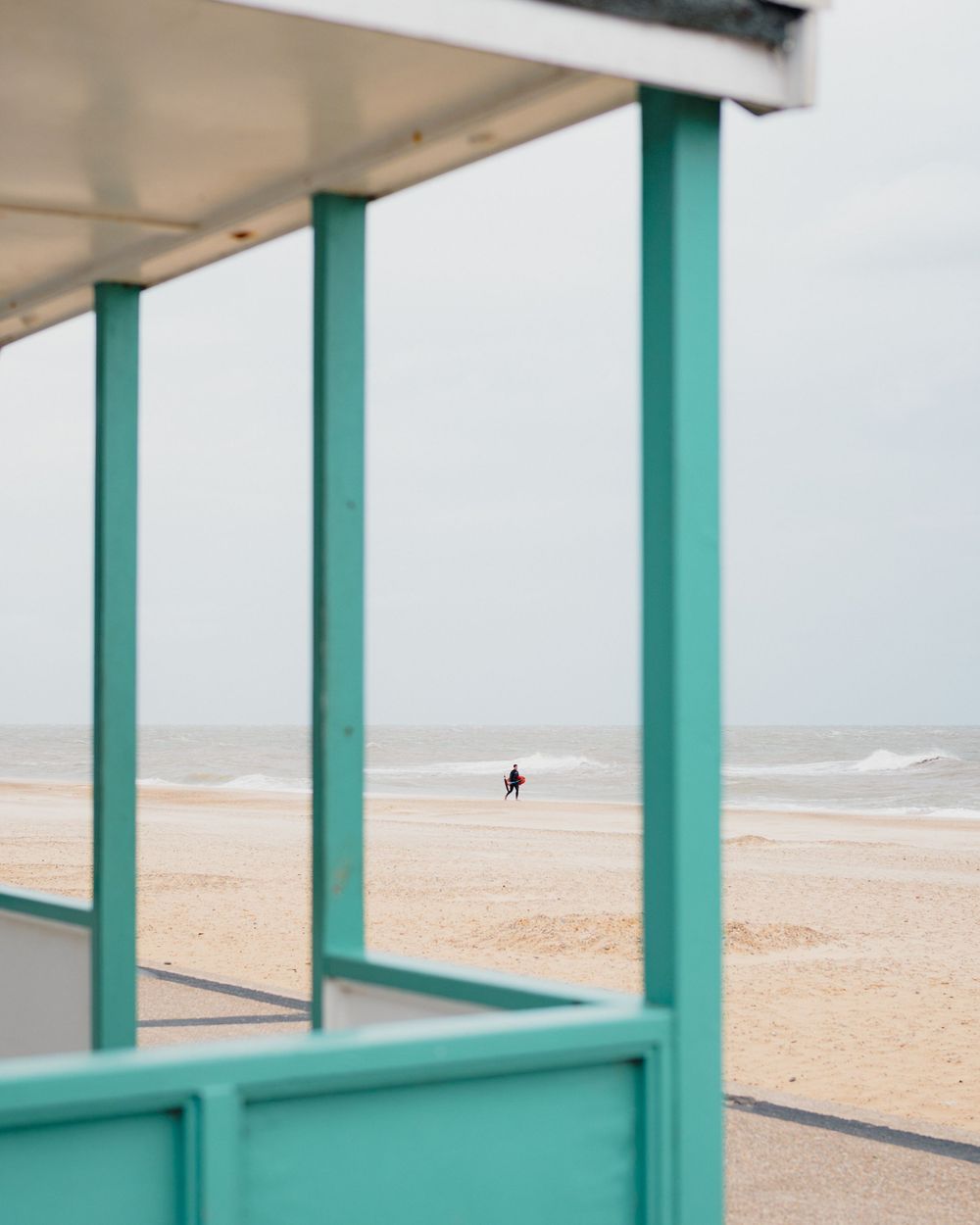 A lone figure walks on a sandy beach viewed through turquoise-framed beach shelter posts on an overcast day.