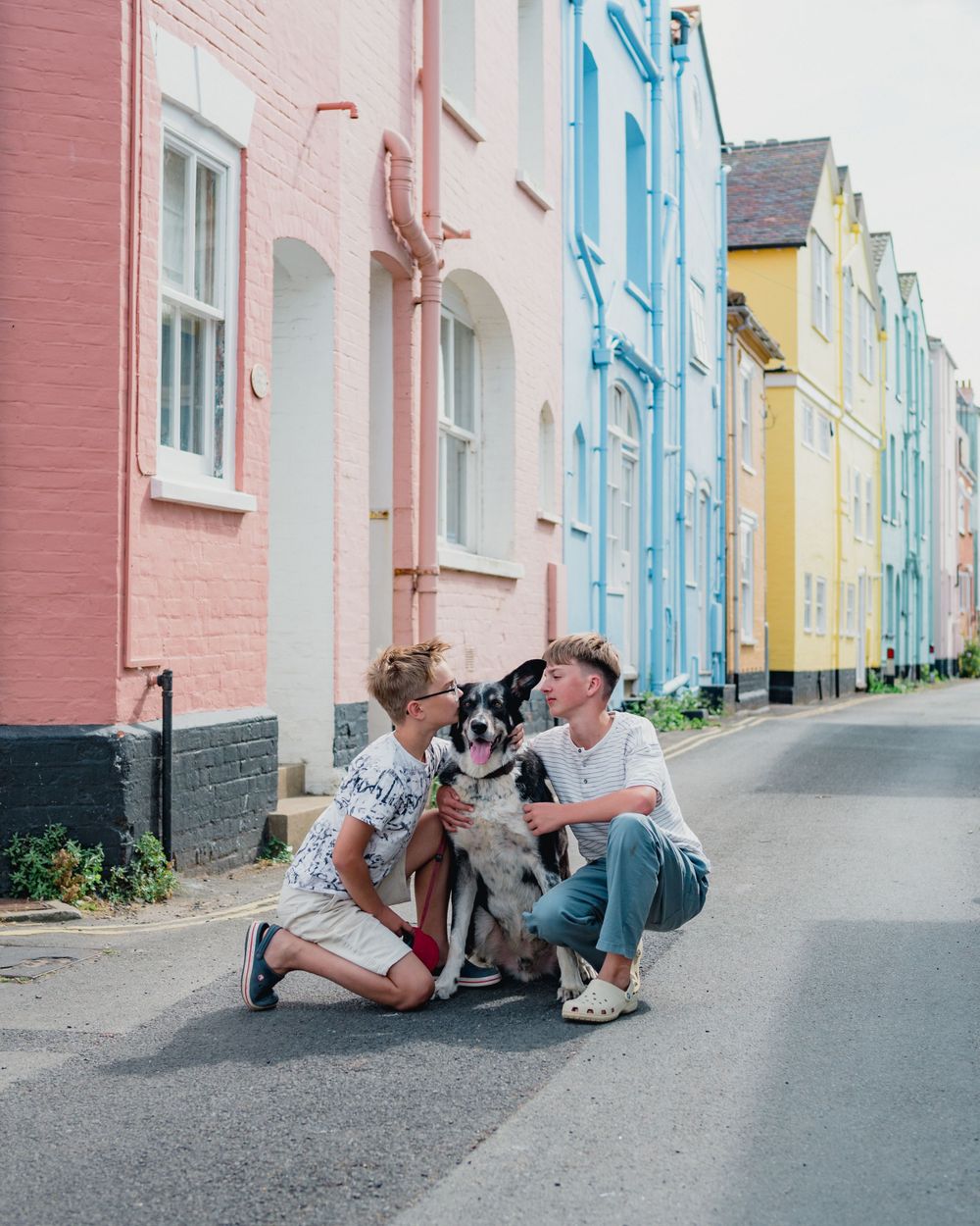 A couple sits and plays with their dog on a charming street lined with colorful pastel houses.