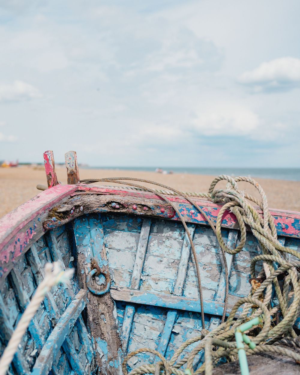 Weathered blue wooden boat hull with ropes and peeling paint against a serene beach backdrop and cloudy sky.