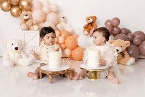 Two babies sit on wooden stools with white cakes during a cake smash photo session with balloons and teddy bears.  Kendall Kuss Photography