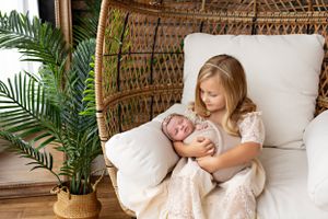 Peaceful scene in wicker chair with tropical plants and white cushions, siblings snuggled up. Newborn photography session with Kendall Kuss