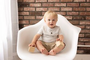 A small child sits in a large white modern chair against an exposed brick wall in a studio setting.