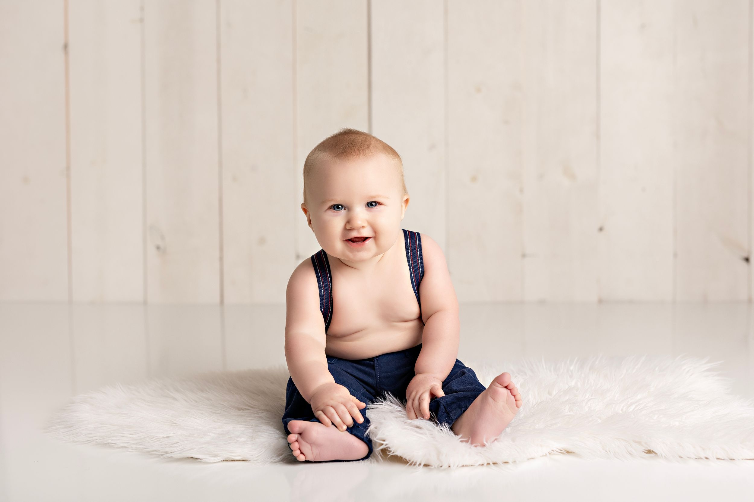 A happy baby wearing navy blue overalls sits on a white furry rug against a light wooden wall background.