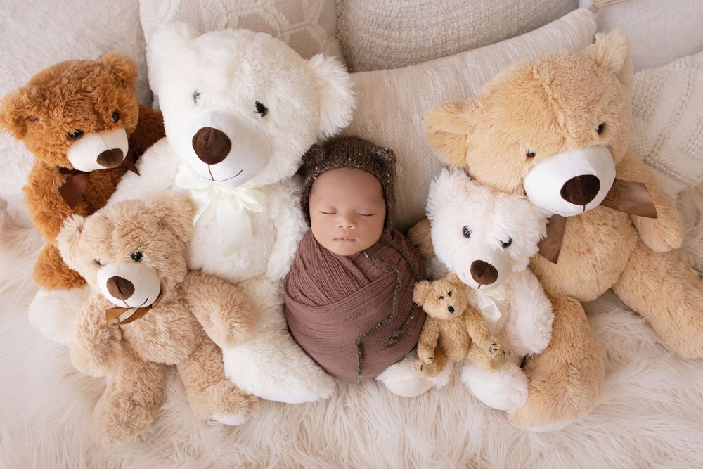 A sleeping newborn wrapped in brown fabric lies surrounded by soft teddy bears on a cream-colored surface.