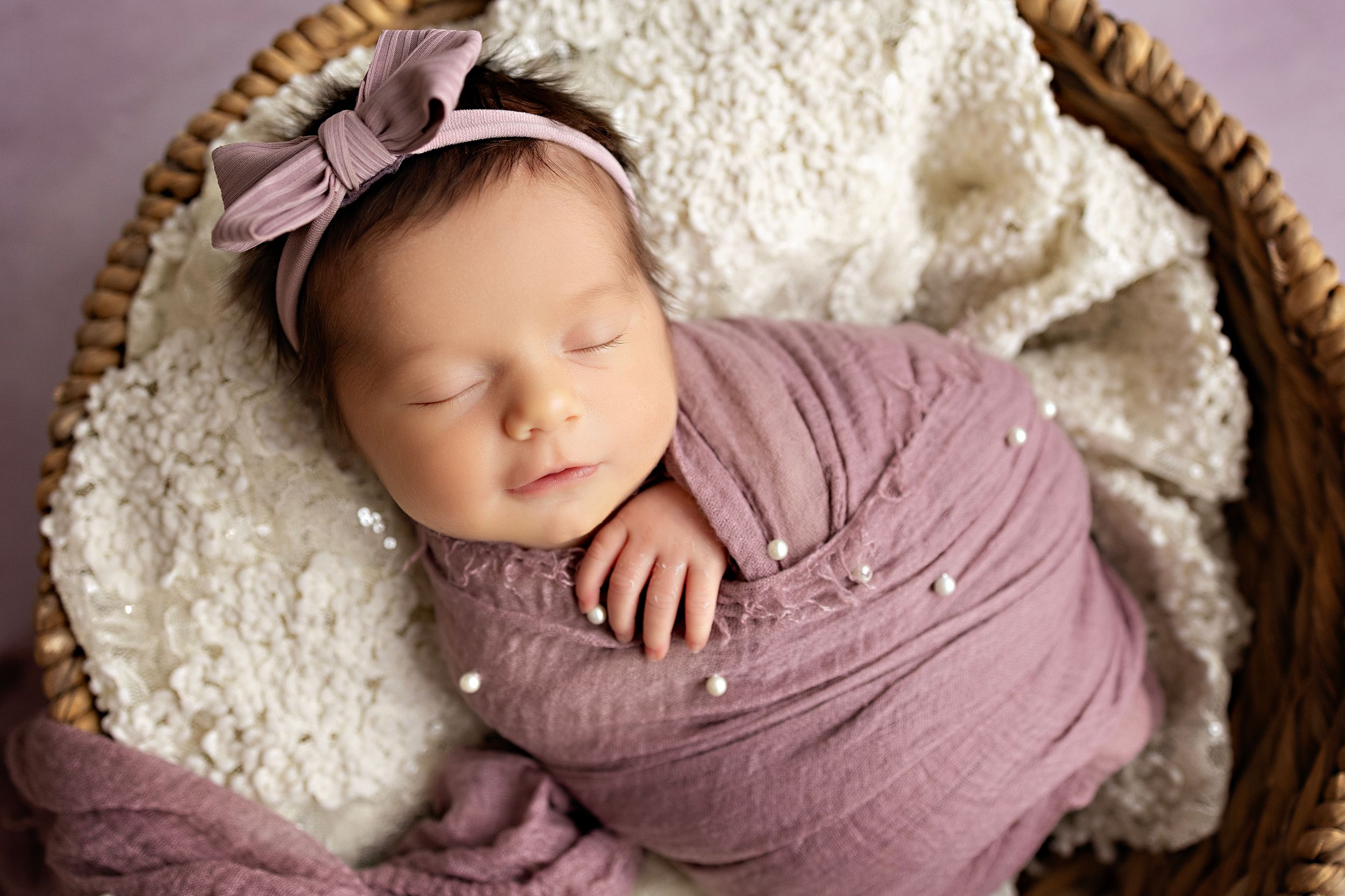 Sleeping newborn wrapped in mauve swaddle with bow headband in woven basket. Newborn photographer Kendall Kuss.