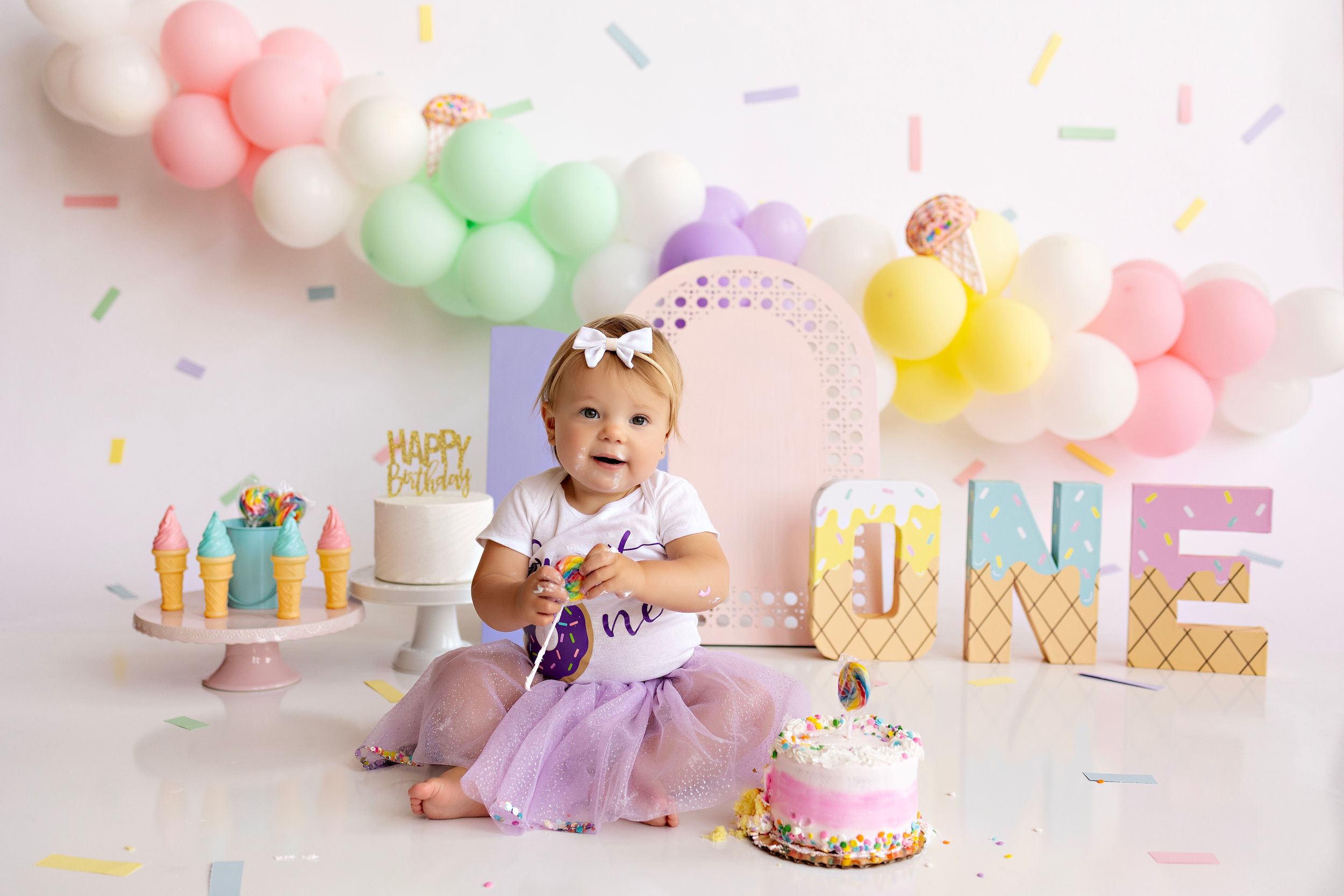 First birthday setup with pastel balloons, cake, and 'ONE' letter display against white backdrop. Kendall Kuss Photography cake smash photos