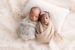 Two sleeping newborn babies swaddled in beige and gray wraps lie peacefully on a white pillow against soft fur.  Kendall Kuss Photography