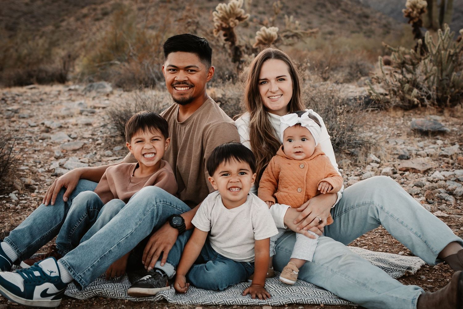 Four people laughing together on a desert trail with rocky mountains and cacti backdrop.