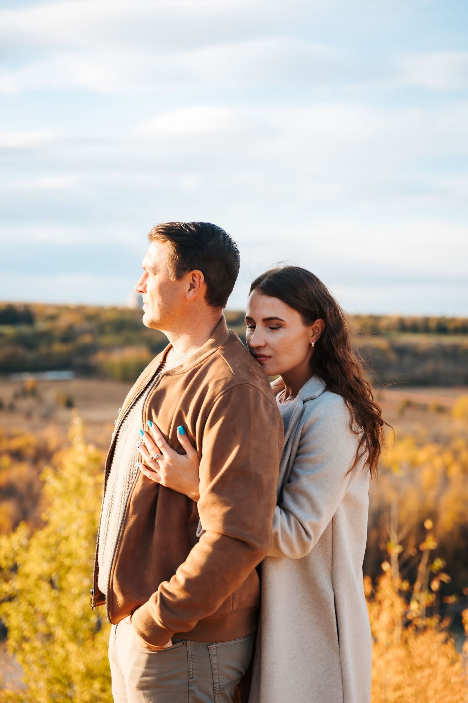 A couple embraces outdoors in autumn, woman resting her head on mans shoulder with golden fall foliage behind them.