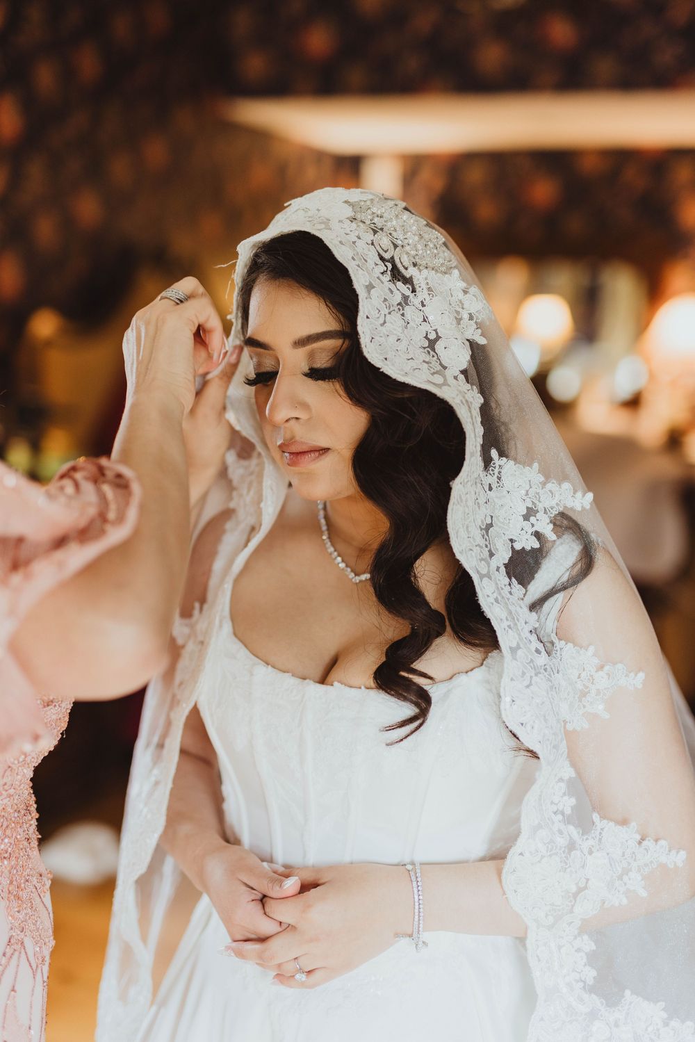 Bride in white lace veil being adjusted by bridesmaid, wearing pearl necklace and bracelet.