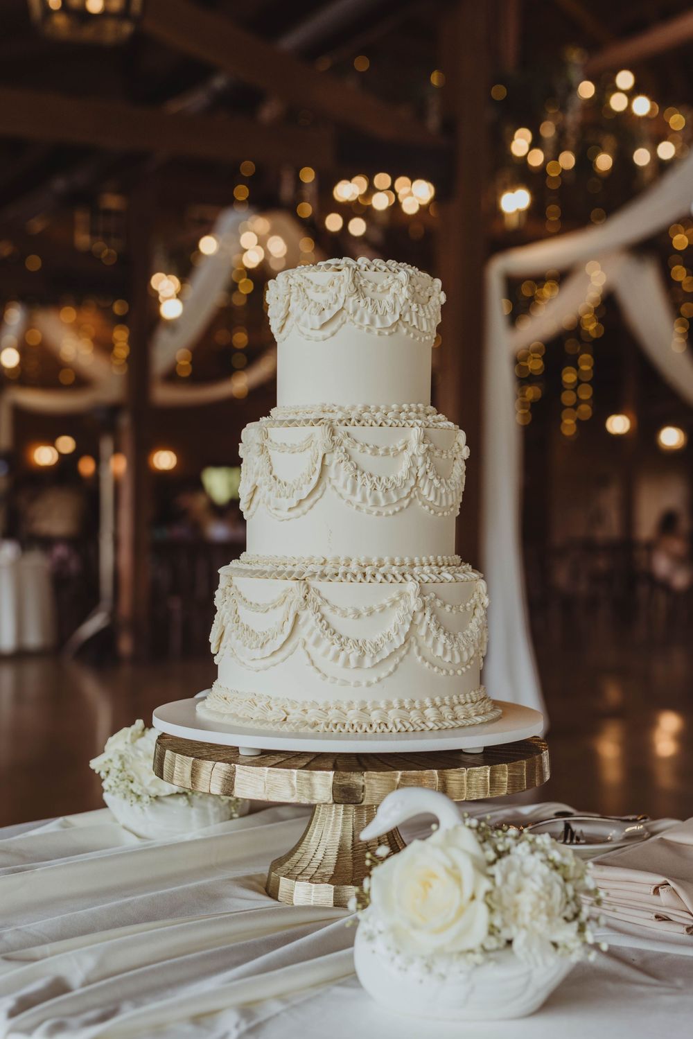 Elegant three-tier white wedding cake with pearl and swag details on a gold stand, set in a warmly lit barn venue.