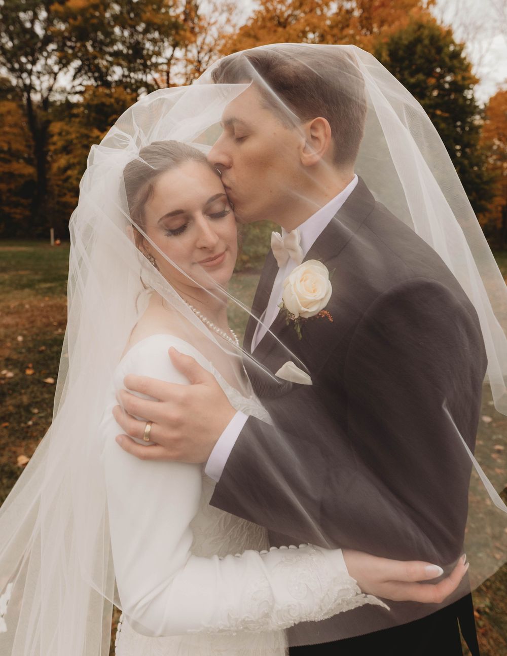 Romantic wedding couple embraces under a flowing veil during an autumn photoshoot.