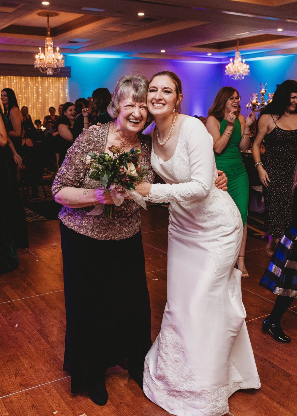 A joyful celebration at a wedding reception with elegant chandeliers, blue and purple lighting, and a wooden dance floor.