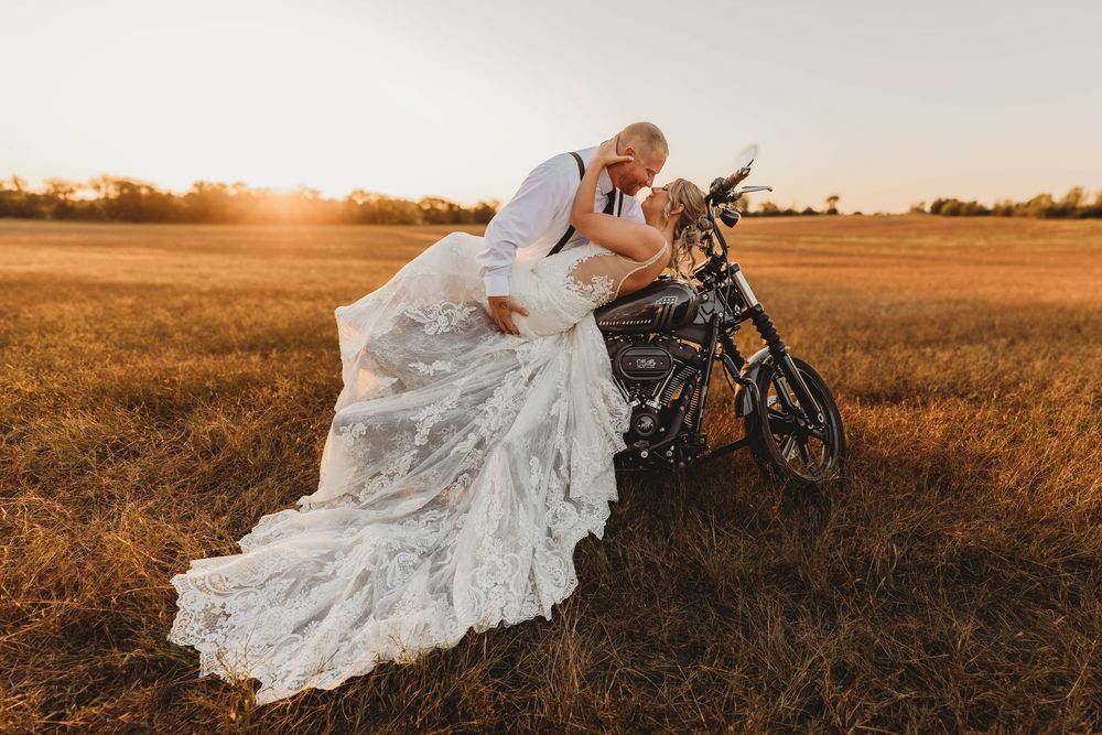 Wedding couple posing with motorcycle in golden field at sunset.