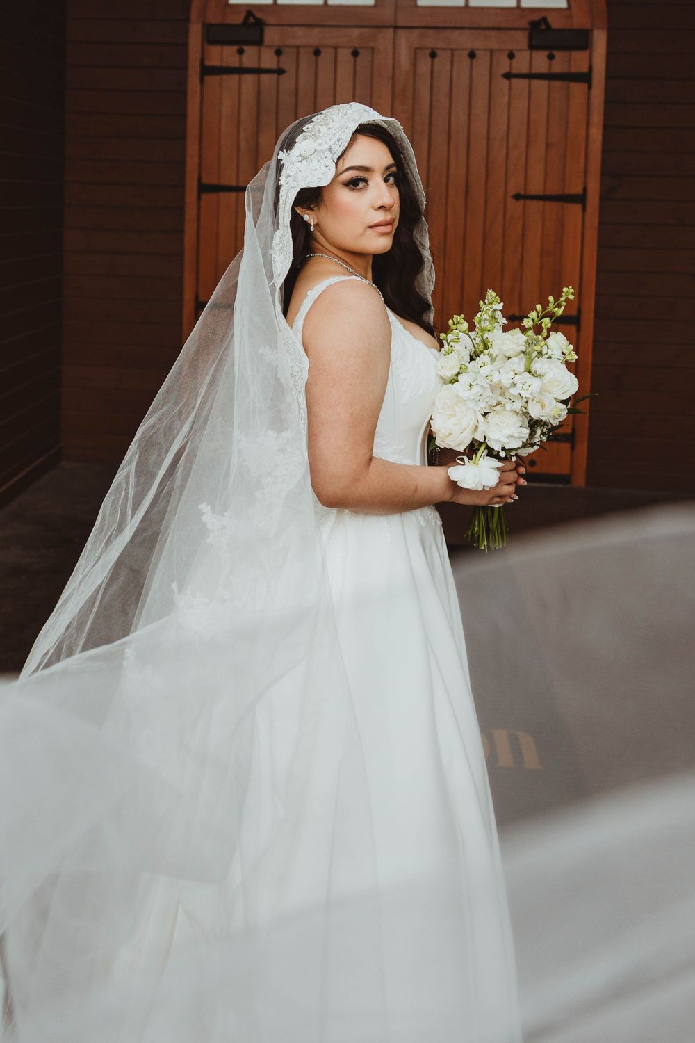 Bride in white A-line gown with lace juliet cap veil holds white bouquet, posing before rustic wooden barn doors.