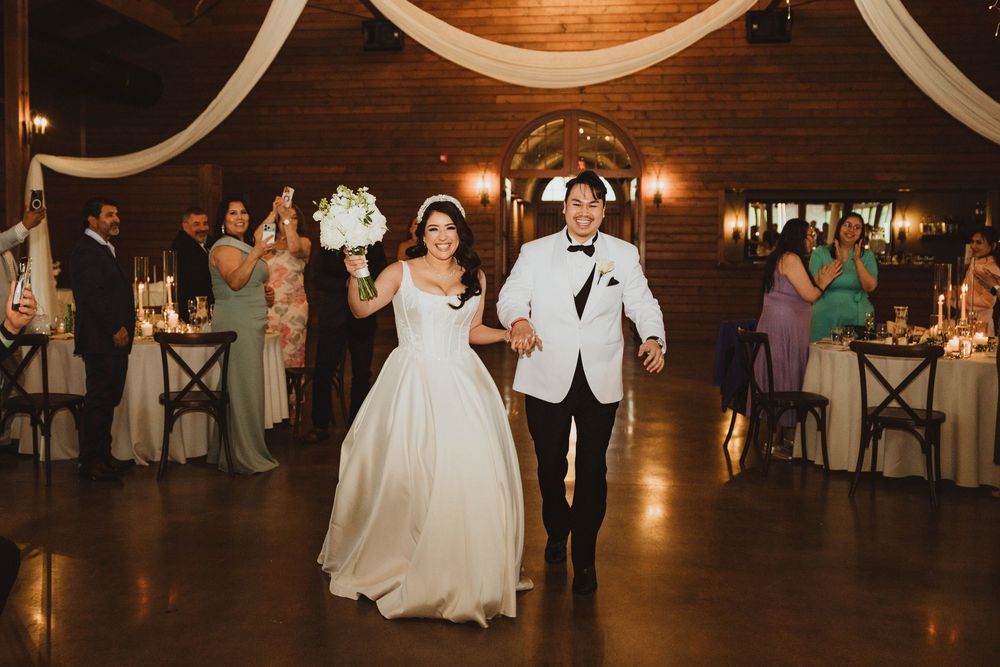 Bride in white gown and groom in white tuxedo make grand entrance at elegant rustic barn wedding reception.