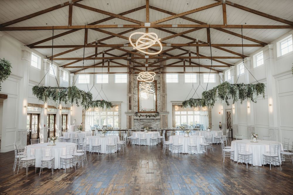 Elegant wedding reception hall with white round tables, hanging greenery, wood beam ceiling, and a modern chandelier centerpiece.