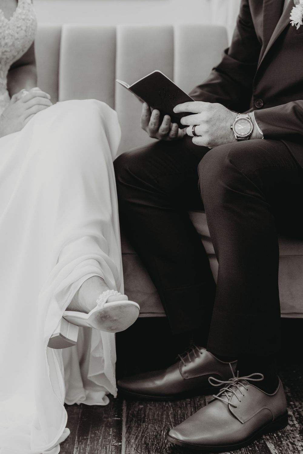 Black and white photo of a bride and groom sitting together, groom reading a book, wearing a suit and dress shoes.