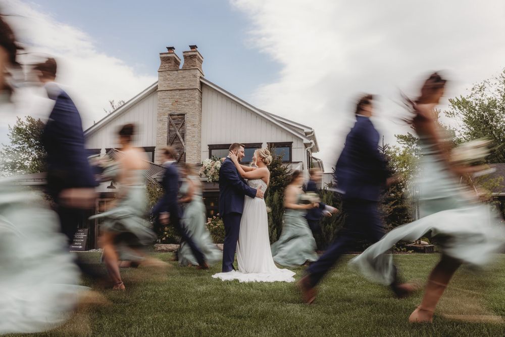 Bride and groom share a kiss while wedding party runs around them in a blur, outside a white barn venue.