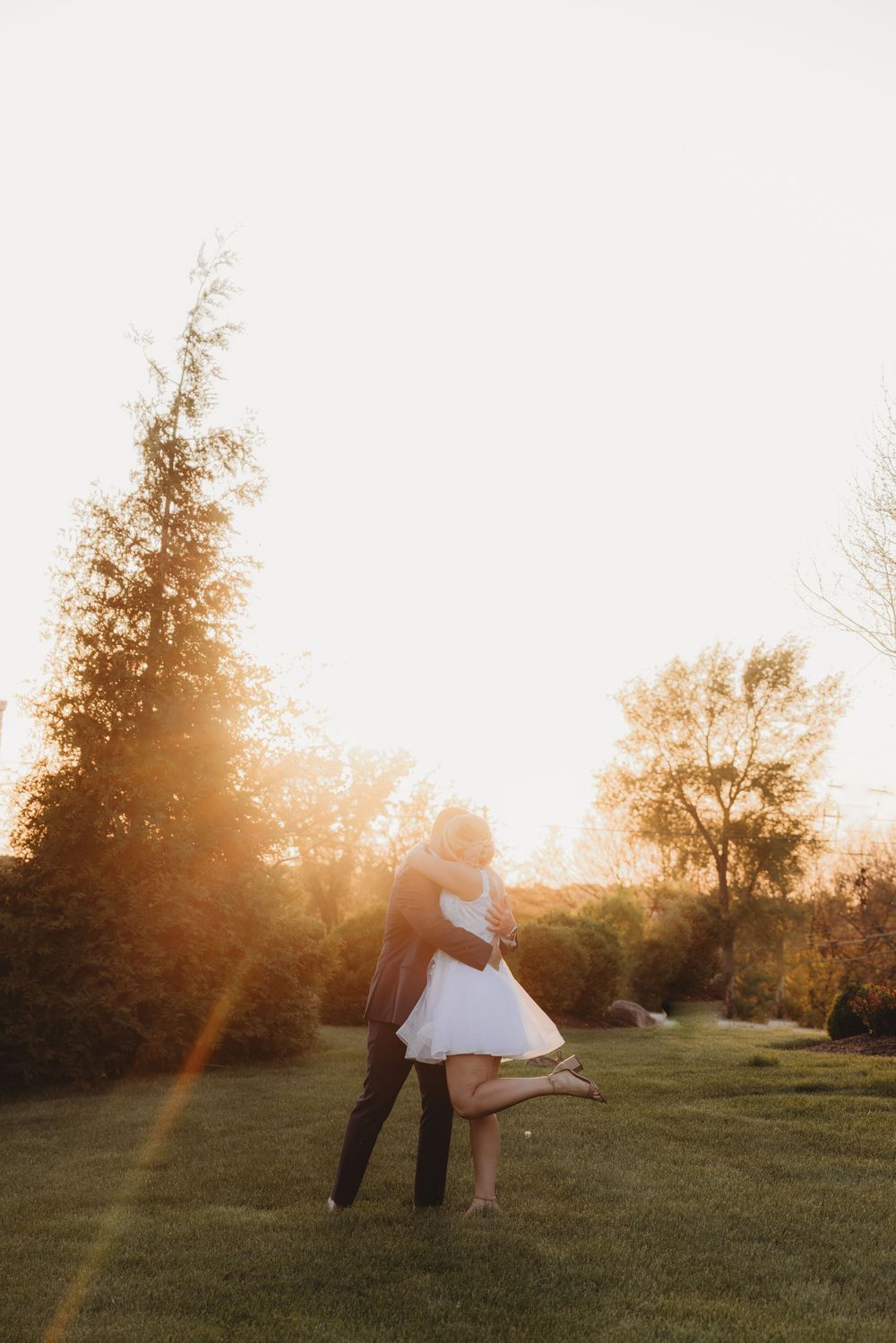 Couple embracing at golden hour sunset in lush green garden, bride in white dress lifted by groom in dark suit.