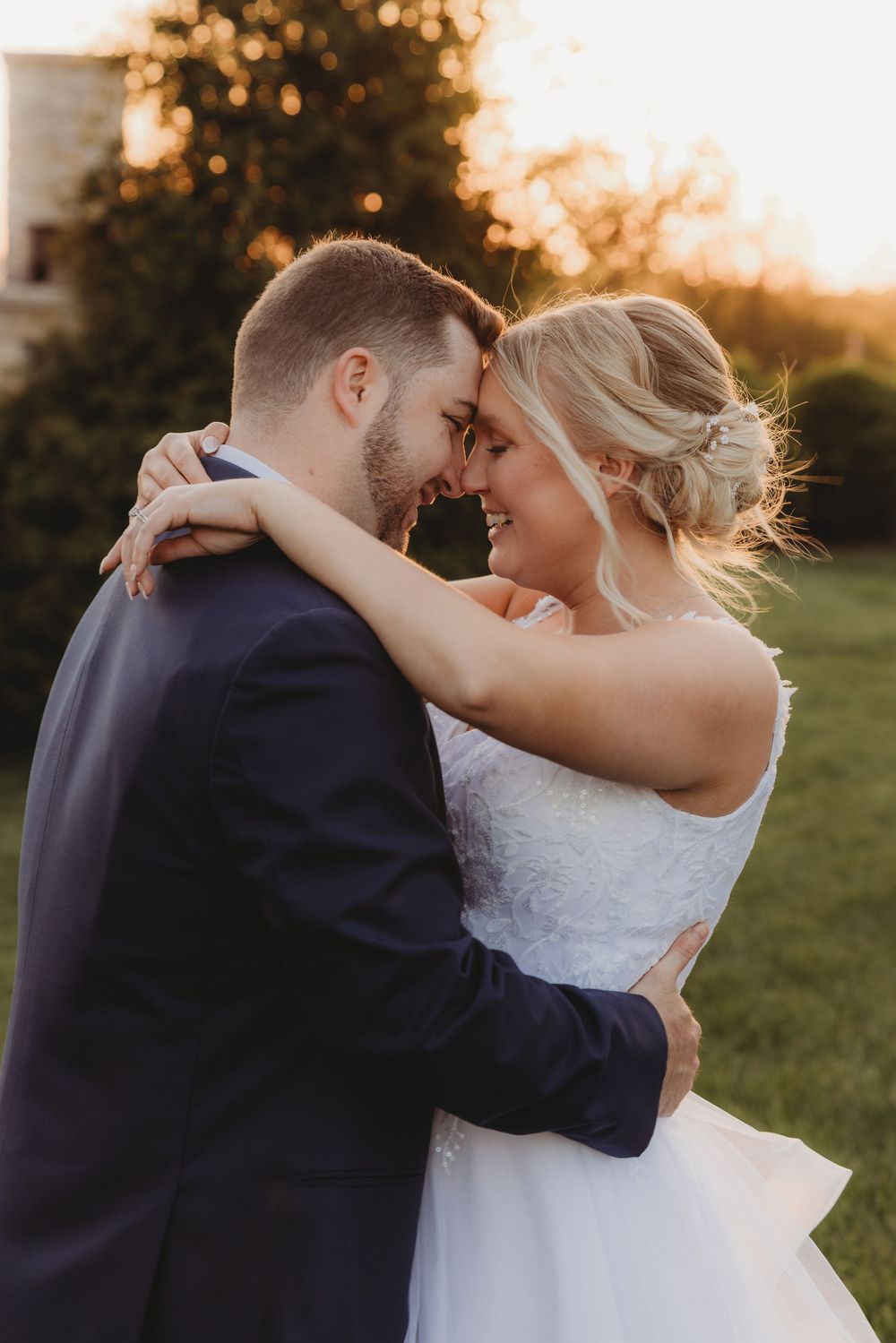 A bride and groom share a romantic forehead touch outdoors at golden hour, she in a white gown, he in a navy suit.