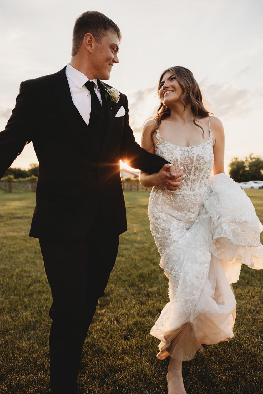 Bride and groom walk hand in hand across green lawn at golden hour, laughing joyfully in elegant wedding attire.