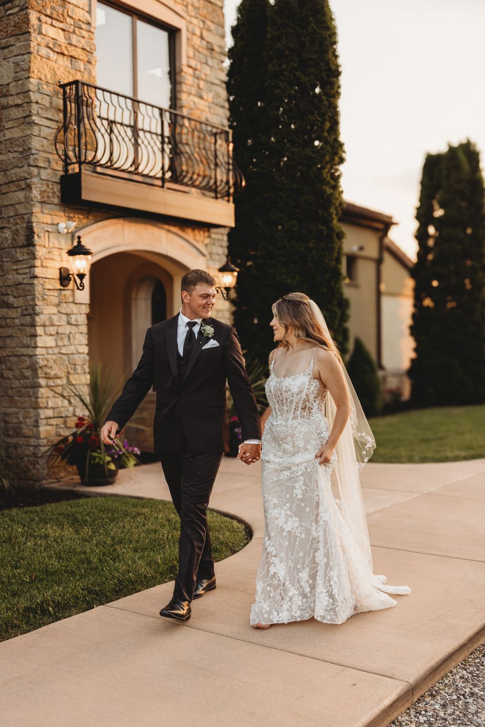 A bride and groom walk hand in hand outside a stone villa at golden hour on their wedding day.