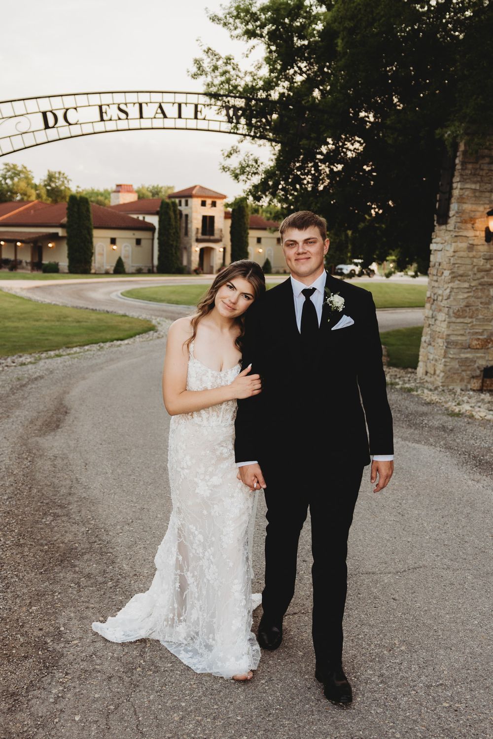 Bride and groom pose together under DC Estate Winery arch at sunset, wearing classic black tuxedo and lace gown.