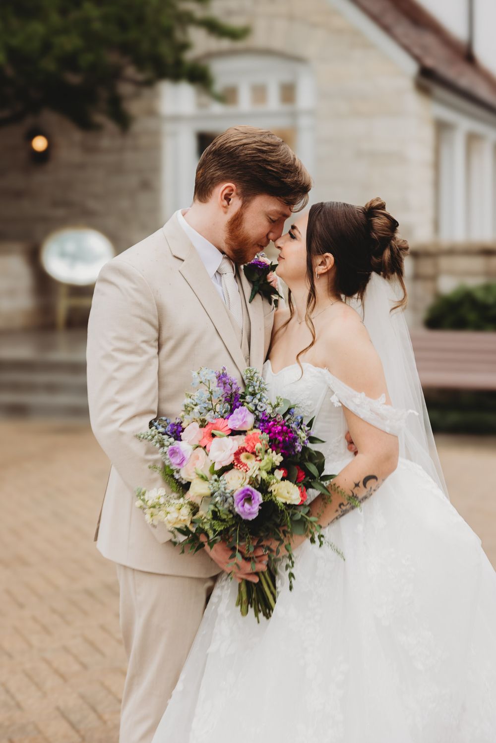 A bride and groom share a romantic moment, holding a colorful bouquet of purple and pink flowers outside a stone building.