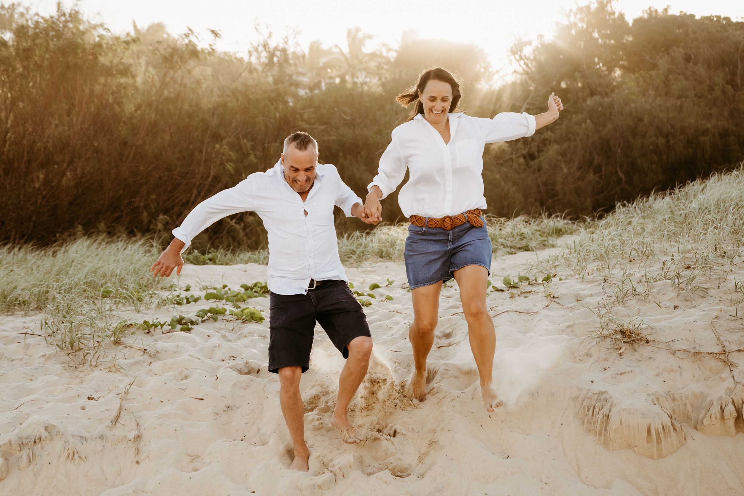 Two people hold hands while playfully jumping on sand dunes at sunset, creating a joyful silhouette against golden light.