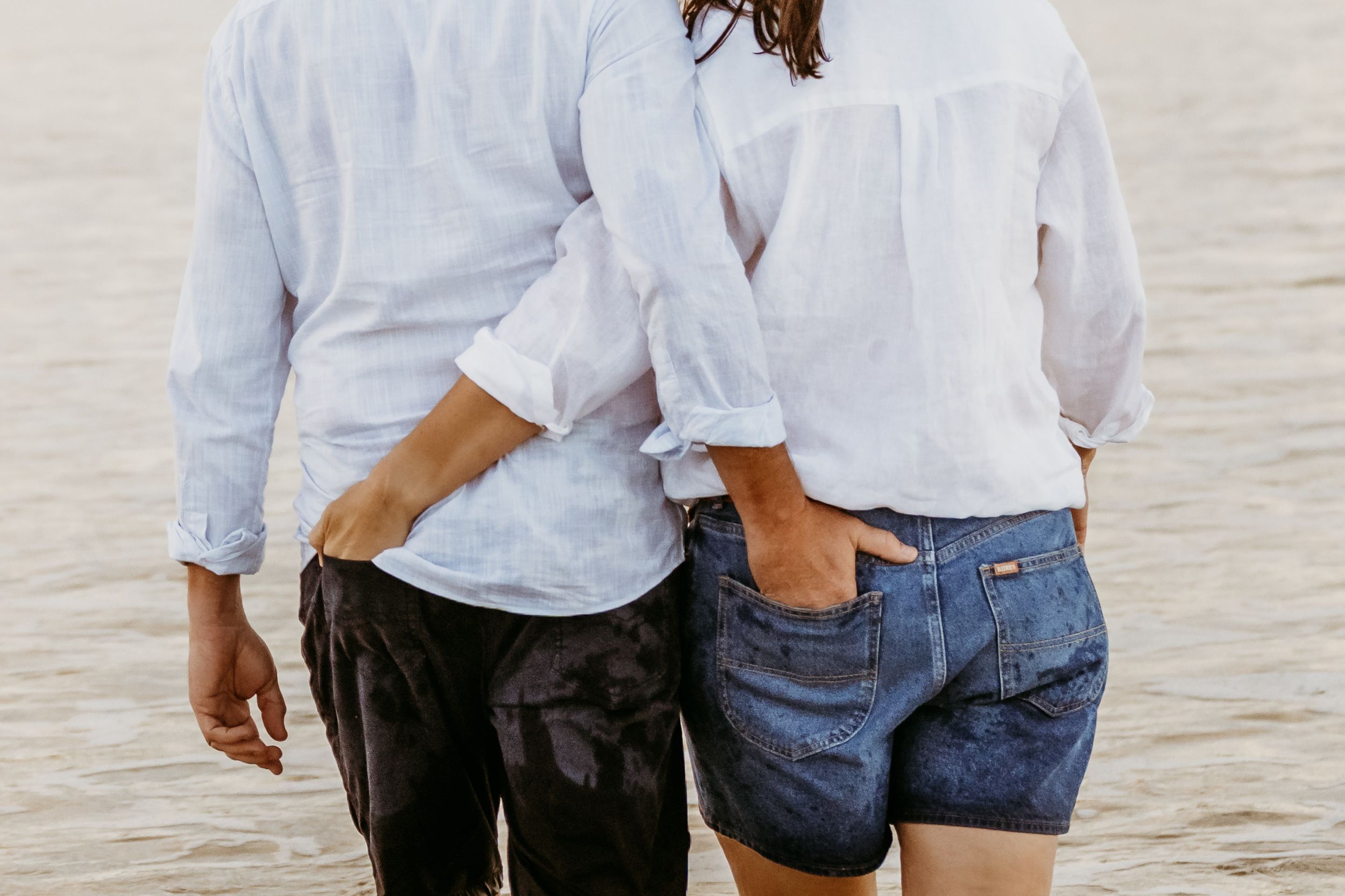 Two people wearing casual white button-down shirts and blue jeans pose together by the waterfront during sunset.